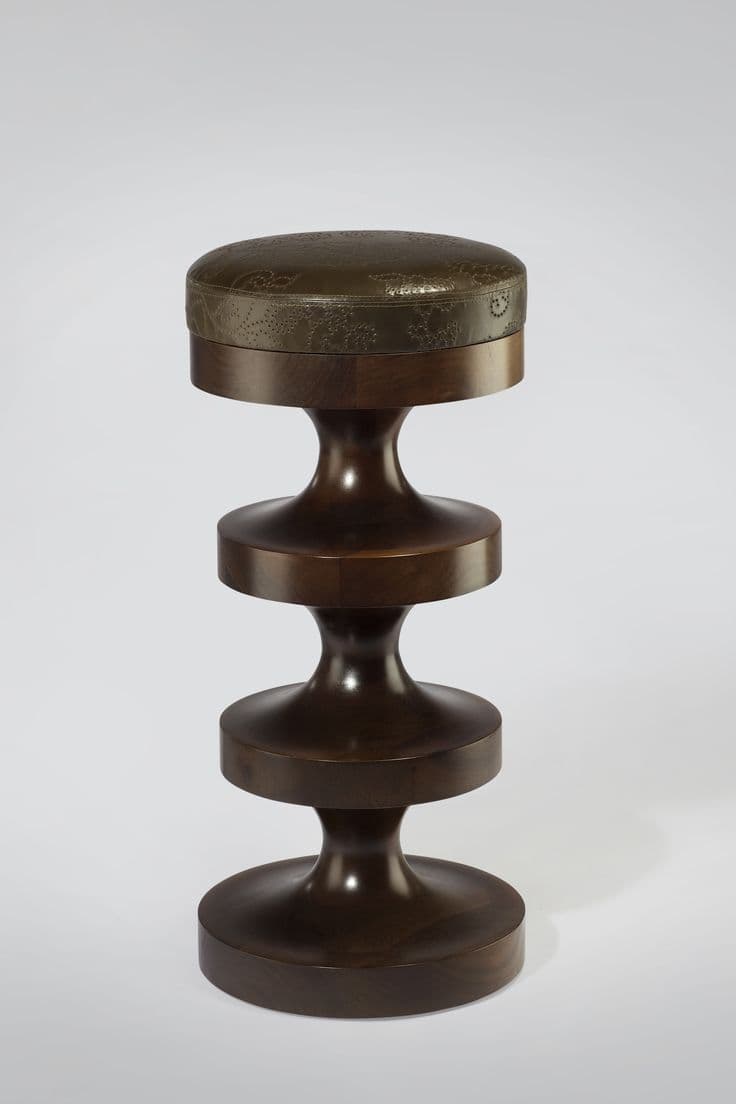 A wooden stool with a padded, upholstered top. The base of the stool is constructed from dark wood, featuring a series of stacked, hourglass-shaped segments. The top is circular and upholstered in a textured material, possibly leather, with an embossed decorative pattern. — 3D model