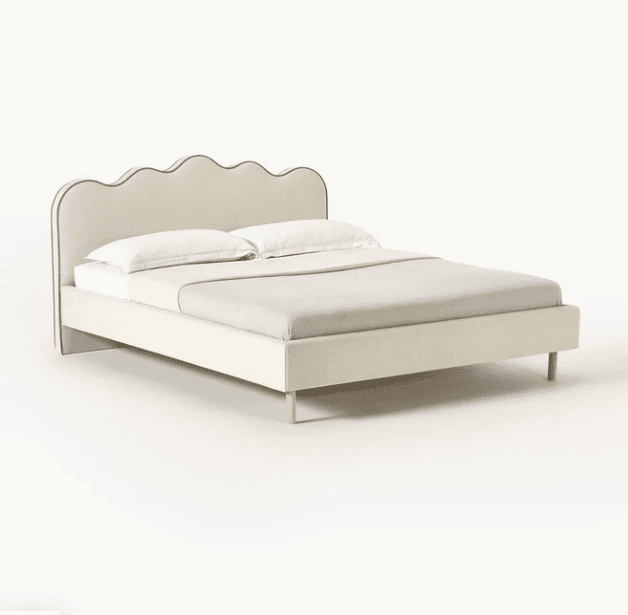 A queen-sized bed frame with a padded headboard and footboard, upholstered in a light beige fabric. The headboard features a distinctive scalloped edge with a darker trim. The bed is supported by four slender, natural wood legs. It is dressed with two white pillows and a light grey blanket. — 3D model