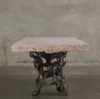 A console table with a rectangular, light pinkish-beige stone top and an intricately sculpted, dark bronze base. The base features an organic, abstract design with interconnected hollow forms and flowing lines, resembling bones or skeletal structures. The stone top has a rough-hewn edge and a textured surface with visible inclusions. — 3D model