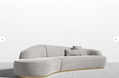 A contemporary curved modular sofa upholstered in light gray velvet, featuring an integrated backrest and armrests, supported by a recessed polished brass plinth base. It includes two matching throw pillows. — 3D model