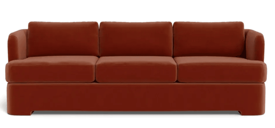 A contemporary three-seater sofa upholstered in a rich terracotta-colored velvet fabric. It features three loose seat cushions and three matching loose back cushions, providing a plush appearance. The arms are gently curved and integrated into the overall frame, flowing seamlessly. The sofa rests on a solid, plinth-like base that extends to the floor, creating a streamlined and low-profile silhouette with a subtle cut-out design at the front bottom edge. — 3D model