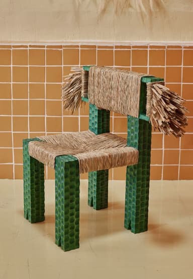 A unique, artisanal accent chair featuring a square, block-like frame crafted from wood with a distinctive deeply textured, almost scalloped or reptilian pattern, finished in a dark green hue. The seat and backrest are constructed from tightly woven natural fiber, likely rattan or raffia, creating a sturdy yet visually appealing surface. The backrest extends slightly beyond the frame, terminating in decorative, untamed fringes of the same natural fiber, adding to its handcrafted aesthetic. The chair's design suggests a blend of rustic charm and contemporary artistic flair. — 3D model