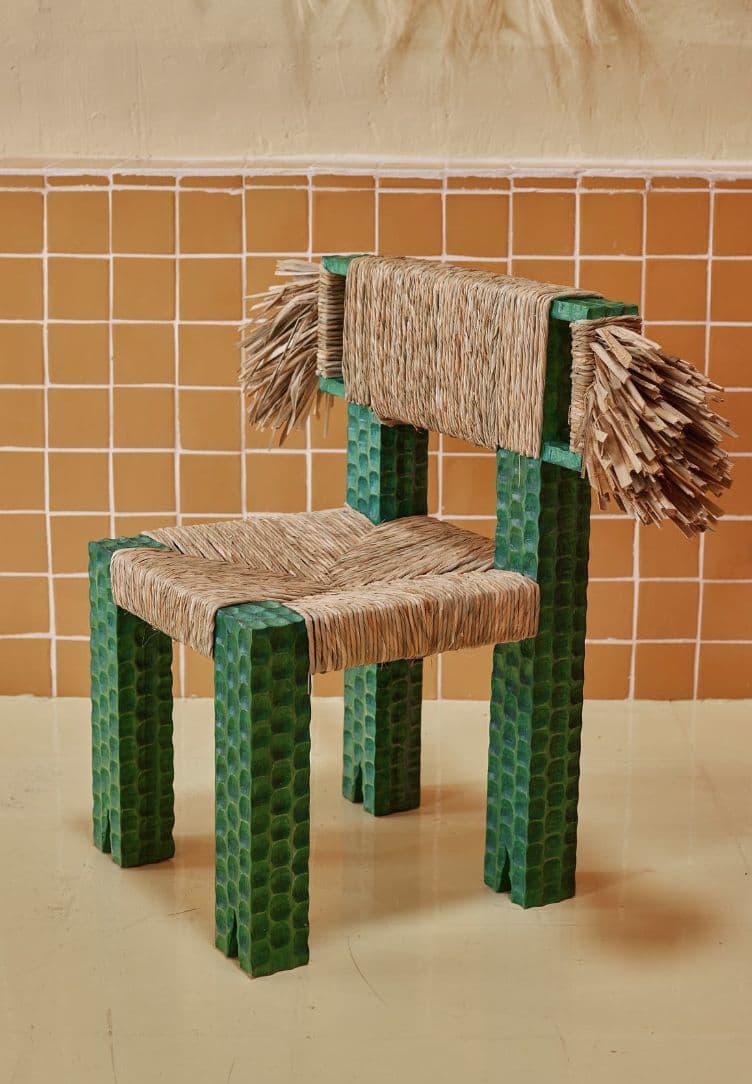 A unique, artisanal accent chair featuring a square, block-like frame crafted from wood with a distinctive deeply textured, almost scalloped or reptilian pattern, finished in a dark green hue. The seat and backrest are constructed from tightly woven natural fiber, likely rattan or raffia, creating a sturdy yet visually appealing surface. The backrest extends slightly beyond the frame, terminating in decorative, untamed fringes of the same natural fiber, adding to its handcrafted aesthetic. The chair's design suggests a blend of rustic charm and contemporary artistic flair. — 3D model