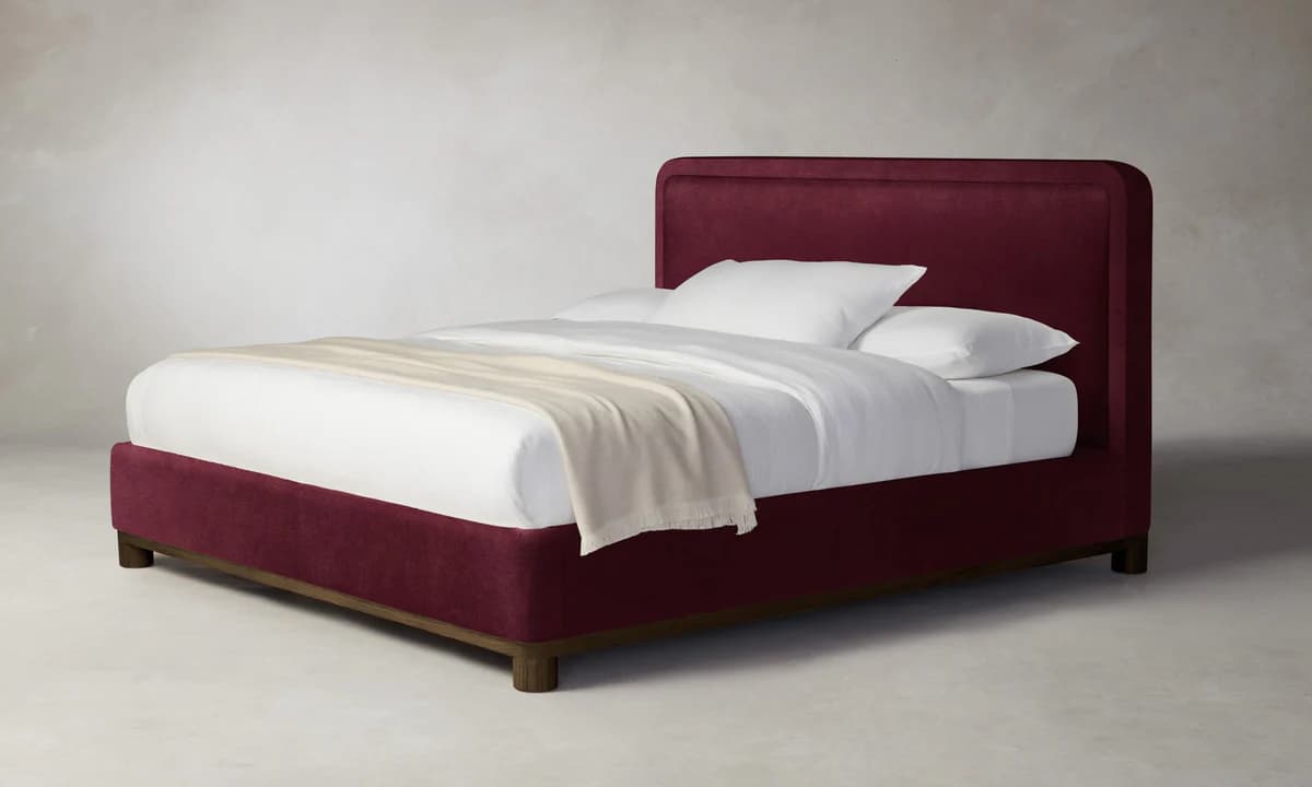 A contemporary upholstered bed frame in a deep red fabric. The bed features a rectangular headboard with slightly rounded top corners and a matching upholstered platform base. It is presented with white bedding, including pillows and a draped throw blanket. The base is elevated by short, dark wooden legs. — 3D model