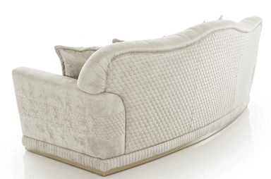 A luxurious three-seater sofa upholstered in light cream or off-white velvet, featuring deep button-tufting across the seat and backrest. The sofa has a gently curved camelback design and rolled armrests adorned with decorative gold-toned metal trim. A pleated or ribbed fabric skirt details the base, which rests on a slender gold plinth. Several matching throw pillows, some with subtle textured patterns, are arranged on the seat. — 3D model