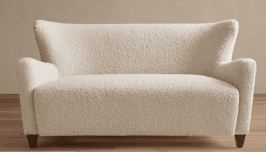 A contemporary two-seater loveseat upholstered in a cream-colored bouclé fabric. It features a high backrest that gently flares outwards at the top, integrated rolled armrests, and a seamless, fully upholstered form. The loveseat is supported by four short, dark, tapered wooden legs. — 3D model