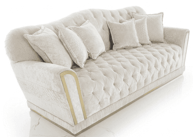 A luxurious three-seater sofa upholstered in light cream or off-white velvet, featuring deep button-tufting across the seat and backrest. The sofa has a gently curved camelback design and rolled armrests adorned with decorative gold-toned metal trim. A pleated or ribbed fabric skirt details the base, which rests on a slender gold plinth. Several matching throw pillows, some with subtle textured patterns, are arranged on the seat. — 3D model