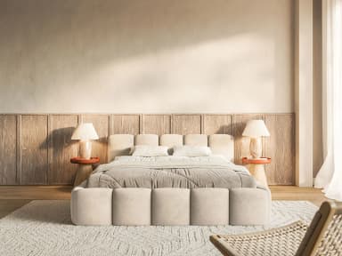 A contemporary platform bed features a plush, segmented design across its headboard and base, creating a soft, inviting aesthetic. The entire frame is upholstered in a light beige fabric, with individual padded sections forming a distinctive visual pattern. The headboard consists of several vertical, rounded panels, providing a comfortable backrest. The low-profile base also showcases similar segmented cushioning around its perimeter. The bed is adorned with white and gray bedding, including throw pillows and a duvet, and is flanked by two small, terracotta-colored side tables, each holding a table lamp with a pleated shade. — 3D model