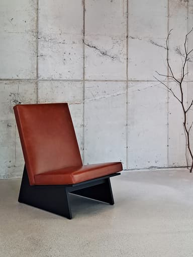 An armless lounge chair featuring a low-profile, angular design. The frame is constructed from black-finished metal, forming a distinctive geometric base. Both the seat and the steeply angled backrest are upholstered in a smooth, warm brown faux leather, providing a contrasting texture and color. The overall aesthetic is minimalist and contemporary. — 3D model