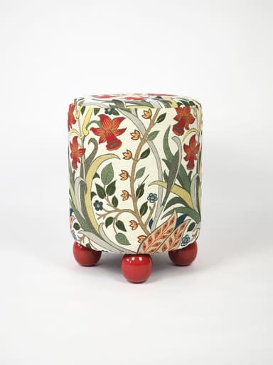 A compact, cylindrical ottoman upholstered in a multi-colored fabric featuring an intricate Arts and Crafts-inspired floral and botanical pattern. The main body of the ottoman rests on four short, spherical, brightly lacquered red feet, providing a contrasting base. The fabric design includes motifs of red flowers, green leaves, and delicate tendrils on an off-white background. — 3D model