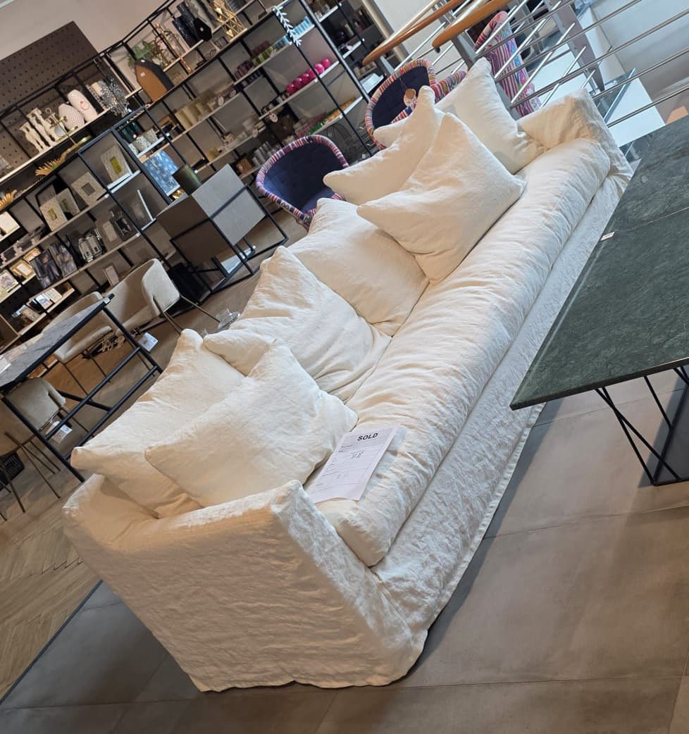 A long, low-profile sofa with a slipcover design. The sofa features plush seat cushions and a generous arrangement of throw pillows in varying sizes. The upholstery appears to be a textured, natural fabric, possibly linen, in an off-white or cream color. The overall aesthetic is relaxed and comfortable, with a slightly wrinkled texture on the slipcover. — 3D model