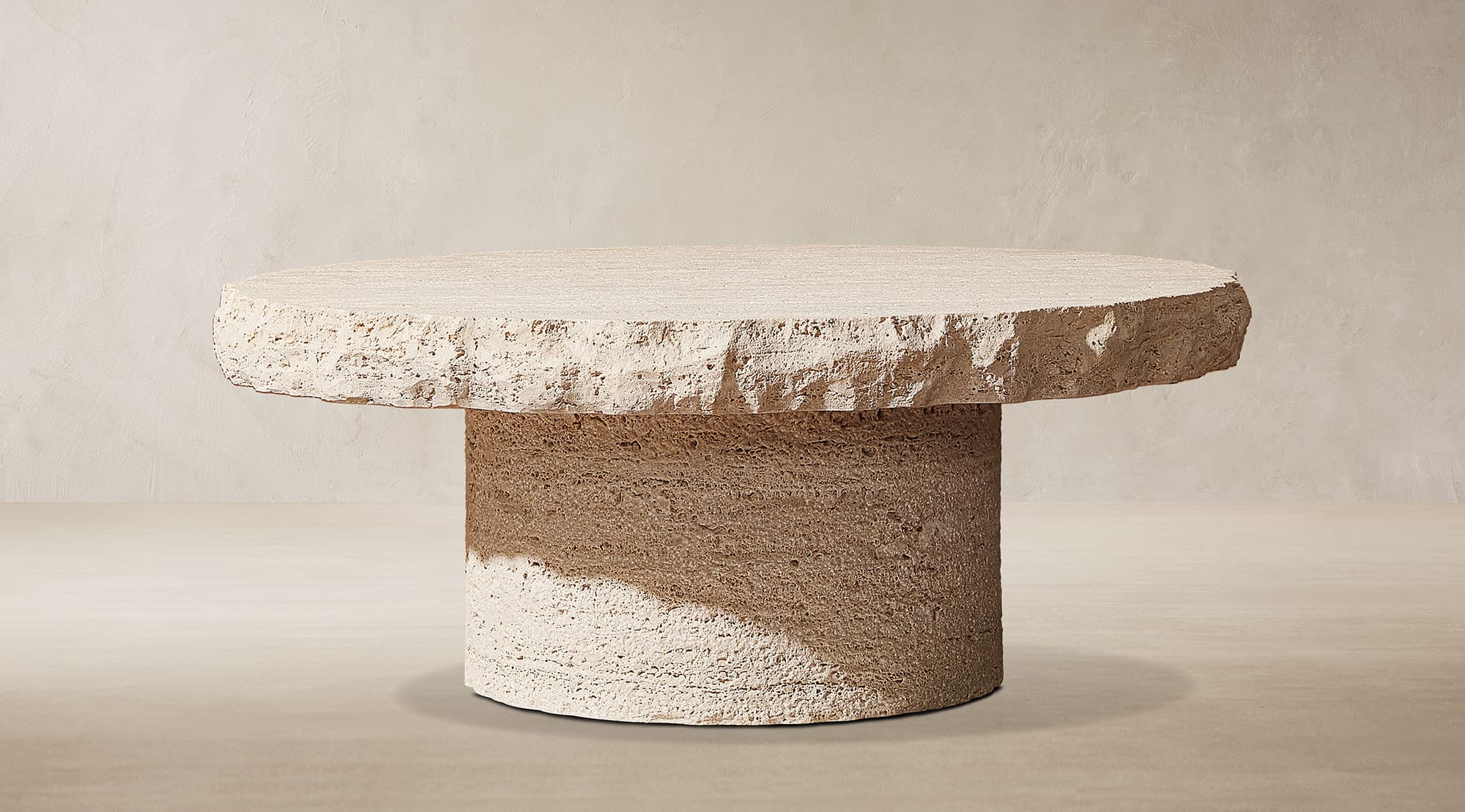 A contemporary round coffee table crafted entirely from rough-hewn travertine stone. The tabletop is a thick, circular slab with natural, unpolished edges, displaying the stone's inherent texture and porosity. It rests upon a robust cylindrical pedestal base, also made from the same textured travertine, creating a cohesive and monolithic appearance. The light beige color and organic form contribute to a raw, earthy aesthetic. — 3D model