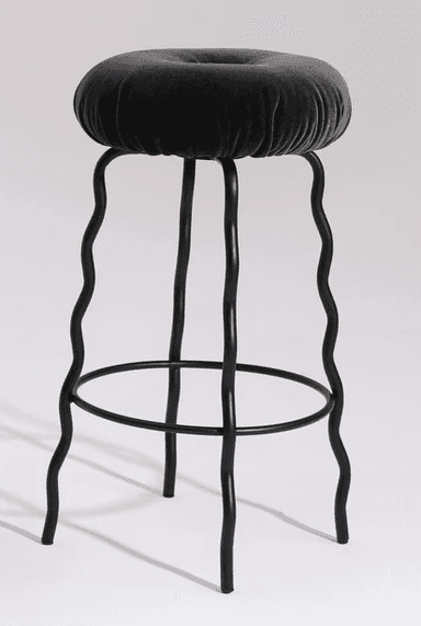 A modern bar stool featuring a deeply cushioned, round seat upholstered in dark grey velvet, distinguished by a central button tuft. The stool's black metal frame consists of four legs, each uniquely designed with a continuous wavy or squiggly profile, extending from beneath the seat to the floor. A circular black metal footrest connects the legs approximately one-third of the way up from the base, providing stability and support. The overall design conveys a playful yet sophisticated contemporary aesthetic. — 3D model