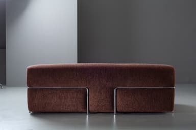 A compact, armless loveseat upholstered in a textured, dark reddish-brown fabric, featuring a minimalist design with a chrome-finished metal bar accent along the base. — 3D model
