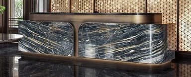 A sophisticated modern reception desk, featuring a substantial body crafted from black and gold veined marble. The desk incorporates rounded corners and two distinct front panels, accented by a metallic bronze-finished frame that wraps around the base and between the marble sections. A raised, L-shaped counter section, also in a dark, possibly stone or metallic finish, sits atop the main structure, supported by the bronze-finished frame. — 3D model