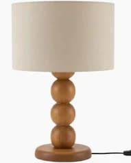 A contemporary table lamp featuring a natural light-toned wood base composed of three stacked spherical elements, culminating in a flat circular pedestal. It is topped with a neutral beige cylindrical fabric lampshade. A black power cord is visible extending from the base. — 3D model