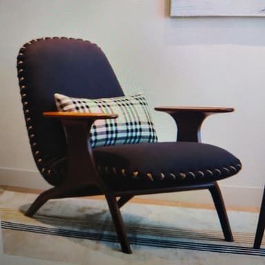 A modern lounge chair featuring a dark wooden frame with integrated, curved armrests and splayed legs. The seat and backrest are upholstered in deep navy blue fabric, accented by prominent exposed whipstitch detailing in a contrasting lighter thread along all edges. A plaid lumbar pillow rests on the seat. — 3D model