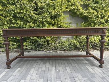 A long, narrow dark wood table, featuring a rectangular top and heavily carved apron and turned legs. The legs are connected by a rectangular stretcher base, presenting a traditional or antique aesthetic. — 3D model