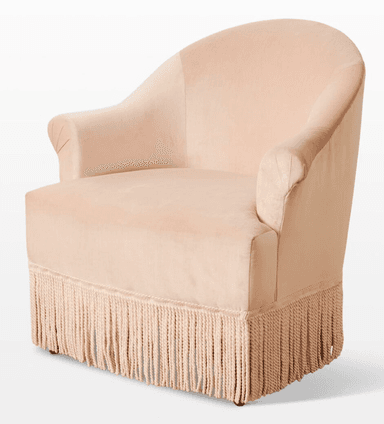 A light pink velvet armchair with a rounded back and arms. It features decorative pleating on the armrests and a prominent fringe along the bottom base. The seat cushion is a lighter, textured fabric. The chair has short, dark wooden legs visible at the very bottom. — 3D model