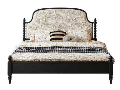 A traditional-style bed featuring a black painted wooden frame with turned posts at all four corners and a footboard. The headboard is upholstered with a light-colored fabric displaying a dense botanical pattern in a darker tone, encased in a curved black wooden frame. The bed is dressed with an assortment of pillows and a duvet, showcasing various patterns including stripes, solids, and a small floral motif, with a black and white checked throw blanket across the foot. — 3D model