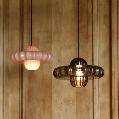 A pale pink pendant light featuring a distinctive ruffled, circular shade. The light hangs from a black cord attached to a cylindrical ceiling canopy. The main shade has a softly undulating or ruffled edge, and a central, raised element with vertical fluting. A diffused light source is visible at the bottom center of the shade. — 3D model