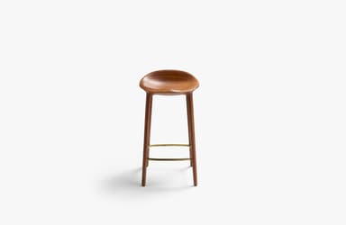 A modern bar stool crafted from dark brown wood, likely walnut, featuring a deeply sculpted saddle seat for ergonomic comfort. It stands on four slender, tapered legs that splay slightly outwards, connected by a triangular brass-toned metal footrest. The design is minimalist and contemporary, highlighting the natural beauty of the wood grain. — 3D model