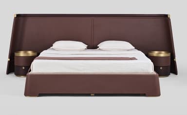 A contemporary upholstered platform bed featuring a broad, angled headboard that extends to either side, integrating cylindrical nightstands. The bed frame, headboard, and nightstands are all upholstered in a dark brown material, likely leather or faux leather. The headboard has subtle paneling and is accented with brass-toned strips and fasteners. The cylindrical nightstands each feature a brass-toned tray top and a lower compartment, possibly with a drawer. The bed is dressed with white bedding and a brown throw blanket. — 3D model