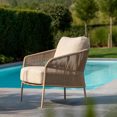 A contemporary lounge chair designed for comfort, featuring a rounded backrest and armrests constructed from tightly woven beige synthetic rope over a minimalist metal frame. It includes a thick, light-colored fabric seat cushion and a matching back cushion. The chair stands on four slender, angled metal legs, coordinating in color with the overall design. — 3D model
