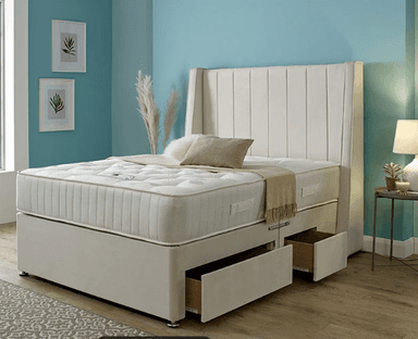 A contemporary upholstered divan bed featuring a tall, winged headboard with vertical channel tufting. The headboard and divan base are covered in a beige fabric. The divan base includes integrated storage drawers on one side and stands on short, chrome-finished feet. A mattress with visible button tufting and piped edges is placed on top of the divan base. — 3D model