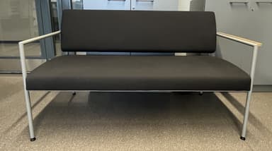 A modern two-seater bench featuring a minimalist design. It has a visible tubular metal frame, black upholstered seat and back cushions, and light-toned wooden armrests. The frame is silver-colored, and the legs are straight with dark footpads. — 3D model