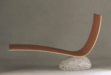 A modern sculptural chaise lounge featuring a gracefully curved, laminated wooden form supported by a single, large, irregularly shaped natural stone base. The wood has a light brown, natural grain finish, and the stone is speckled grey, creating a striking contrast of organic materials and minimalist design. — 3D model