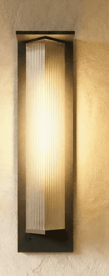 A tall, rectangular wall-mounted light fixture featuring a dark-toned metal backplate and a prominent fluted glass diffuser. The glass section is ribbed vertically and appears to be illuminated from within, casting a warm glow. The top of the metal frame and glass diffuser show an angular, stepped design, giving it a distinct Art Deco or classic modern aesthetic. The light is shown mounted on a textured, light-colored wall. — 3D model