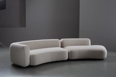 A two-piece modular sofa featuring a distinct curved silhouette and upholstered in a light-colored, textured boucle fabric. The design is contemporary with rounded edges and a low profile, comprising a left arm section and a right armless chaise-like section that together form a gentle curve. The sofa sits close to the floor on concealed or minimalist feet. — 3D model