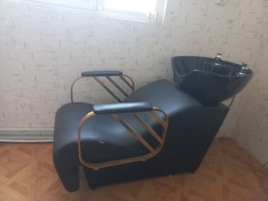 A modern salon shampoo chair featuring a reclined seating position and an integrated black ceramic basin at the rear. The chair is upholstered in black material, likely faux leather or vinyl, with a sleek, curved design. It includes gold-finished metal armrests with padded black tops, complementing the overall contemporary aesthetic. — 3D model