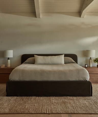 A modern upholstered bed featuring a low-profile platform base and a gently curved headboard, all covered in a dark brown fabric. The bed is dressed with light beige/tan bedding, including a duvet, two standard pillows, and a long bolster pillow. The design is minimalist and contemporary. — 3D model