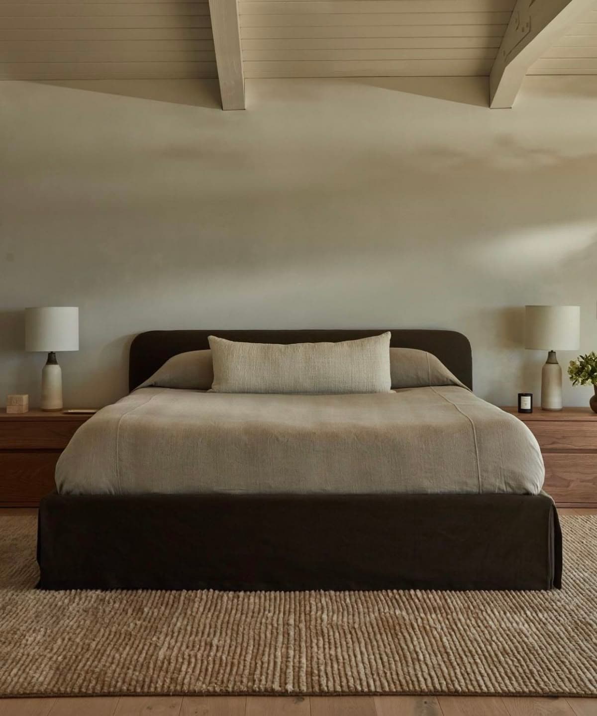 A modern upholstered bed featuring a low-profile platform base and a gently curved headboard, all covered in a dark brown fabric. The bed is dressed with light beige/tan bedding, including a duvet, two standard pillows, and a long bolster pillow. The design is minimalist and contemporary. — 3D model