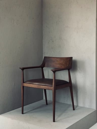 A modern wooden dining chair featuring a curved backrest, integrated armrests, and an upholstered leather seat, all supported by slender, tapered legs. — 3D model