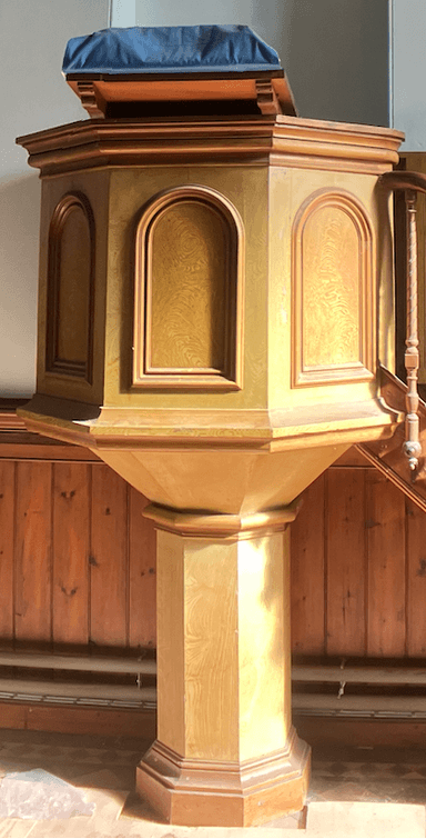 A light-toned wooden hexagonal pedestal featuring an octagonal display top. The upper section of the pedestal is hexagonal with six distinct panels, each adorned with a raised arched inset design. It is supported by a central, tapered octagonal column which transitions into a broad, stepped hexagonal base. The piece is constructed with visible wood grain and includes darker wood trim accents, indicative of traditional craftsmanship. — 3D model