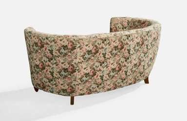 A vintage-style, curved sofa upholstered in a floral fabric with a pastel color palette. The sofa features a rounded backrest with vertical channeling and integrated armrests that curve inwards. It is supported by four short, tapered wooden legs. — 3D model