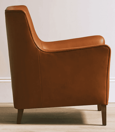 A contemporary armchair upholstered in cognac-colored leather, featuring a broad seat, low-profile rolled armrests, and a gently angled backrest. The chair is supported by four tapered wooden legs, visible beneath its substantial body. Visible stitching defines the edges and seams of the upholstery, contributing to its tailored appearance. — 3D model