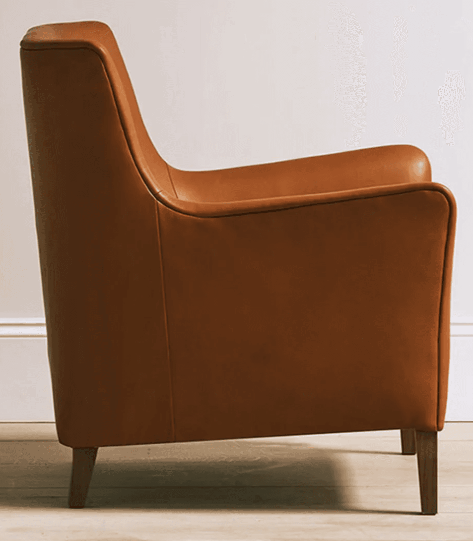 A contemporary armchair upholstered in cognac-colored leather, featuring a broad seat, low-profile rolled armrests, and a gently angled backrest. The chair is supported by four tapered wooden legs, visible beneath its substantial body. Visible stitching defines the edges and seams of the upholstery, contributing to its tailored appearance. — 3D model