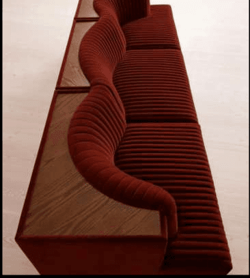 A long, contemporary modular seating unit featuring deep red channel-tufted velvet upholstery. It has a distinct long, curved backrest which transitions into an integrated wooden shelf or table surface. The seat cushions are also channel-tufted, providing a ribbed texture. The overall form suggests a lounge or public seating application, combining comfort with functional surface space. — 3D model