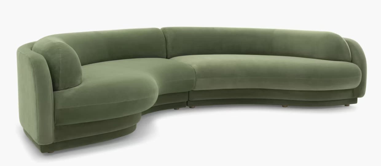 A modular, curved sectional sofa upholstered in a soft, green velvet-like fabric. The sofa features rounded edges and a layered, almost sculptural base. The design is contemporary with organic, flowing lines, suggesting a modern or art deco-inspired aesthetic. It appears to be composed of at least two connected sections to form the corner shape. — 3D model