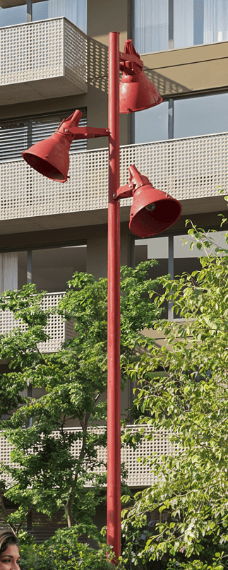 A tall, slender pole supporting three adjustable cone-shaped lamps arranged vertically. The lamps are oriented outwards and downwards, each featuring a wide, flared metal shade with an exposed bulb socket. The pole and lamp fixtures are a uniform, vibrant red color, suggesting a durable metal construction suitable for outdoor or industrial settings. The joints at the lamps allow for articulation, enabling directional lighting. — 3D model