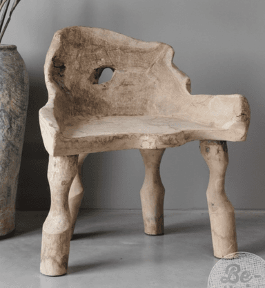 A unique lounge chair carved from a single piece of weathered, light-colored wood, likely teak or a similar robust timber. It features an organic, sculptural form with integrated backrest and armrests, showcasing natural imperfections and grain patterns. The legs are irregularly shaped, contributing to its primitive and rustic aesthetic, and there is a distinct, naturally occurring or carved hole in the upper backrest. — 3D model