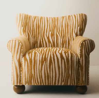 A compact upholstered armchair featuring a distinctive gold and off-white zebra-print fabric. It has a high, subtly winged backrest and classic rolled armrests. The chair is supported by four short, dark wooden bun feet, and shows visible piping detail along its seams. — 3D model