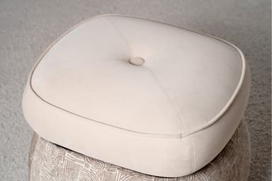A two-tiered ottoman with a rounded, tufted top cushion and a patterned base. The top cushion is upholstered in a smooth, light beige fabric and features a single button tuft in the center, outlined by stitched seams. A contrasting piping trim defines the edge of the top cushion. The base is wider than the top and is upholstered in a textured fabric with a repeating abstract pattern in shades of beige and white, resembling arcs and linear shapes. The ottoman is situated on a textured, light-colored carpet. — 3D model