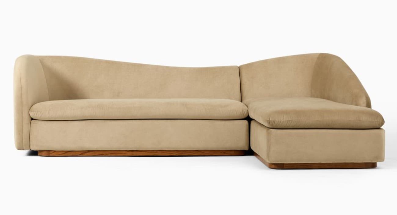 A contemporary curved sectional sofa upholstered in a light beige, plush fabric, featuring an integrated right-facing chaise lounge and a recessed wooden plinth base. — 3D model