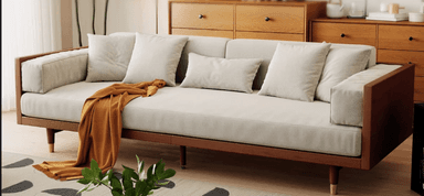 A modern sofa featuring an exposed dark wood frame that encompasses the seat and back, providing structural and aesthetic definition. The sofa is upholstered in a light beige or cream fabric, consisting of three main seat cushions and multiple back and throw pillows, offering a plush seating experience. The frame rests on slender, round tapered legs, which are dark wood at the top and transition to a metallic, possibly brass or gold-finished, cap at the bottom, adding a touch of elegance. An orange throw blanket is casually draped over one side of the sofa. — 3D model
