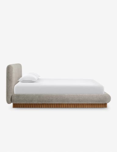 A side profile view of a modern upholstered bed with a curved headboard and a wooden slatted base. The bed frame is upholstered in a textured, light-colored fabric, and it features rounded edges. A white mattress with white bedding and a single white pillow are visible on top. — 3D model